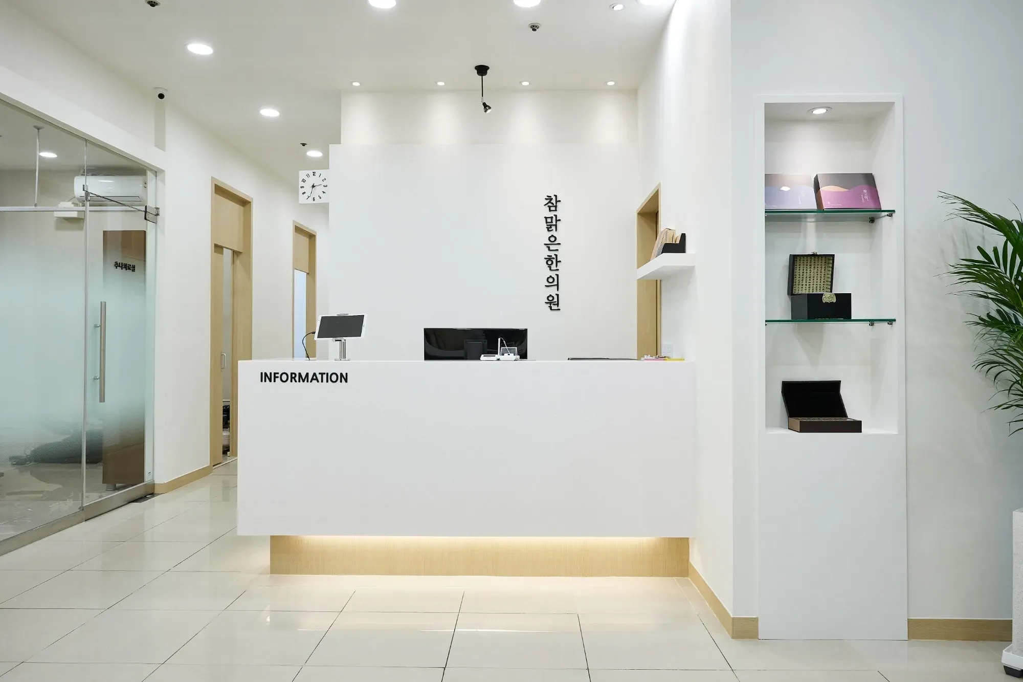 INFORMATION 접수대 뒤 흰 벽에 참맑은한의원 이름이 세로로 걸린 입구, 오른쪽 벽에 유리 진열장
